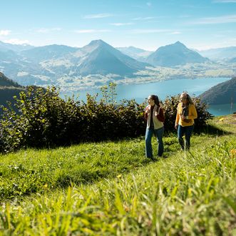 Zwei Frauen gehen auf einer grünen Wiese mit Blick auf einen See und Berge wandern, im Vordergrund hohes Gras und Sträucher.