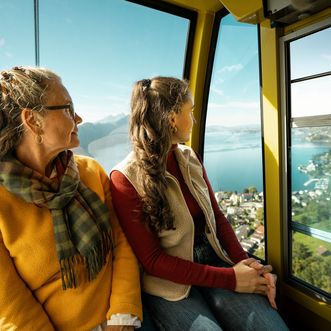 Eine Frau und ein junges Mädchen sitzen in einer Gondel und blicken nach draussen auf den See und die Berge. Durch die Fenster ist eine sonnige Landschaft mit Wasser, Hängen und einem Ort am Ufer zu sehen.
