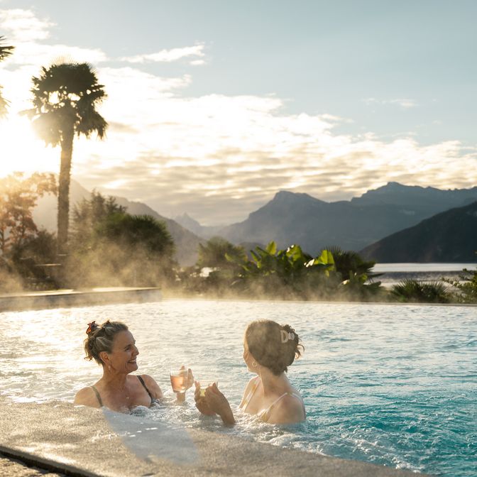 Zwei Frauen stehen in einem Aussenpool und halten Getränke in der Hand, während im Hintergrund Dampf über dem Wasser aufsteigt, Palmen, Berge und ein See im warmen Abendlicht zu sehen sind.