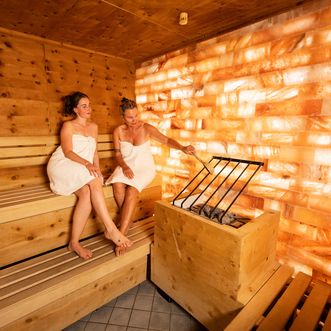 Zwei Frauen sitzen in weisse Badetücher gewickelt auf einer Holzbank in einer Sauna. Eine von ihnen hält eine Schöpfkelle über einen mit Steinen gefüllten Ofen, daneben leuchtet eine Wand aus orangefarbenem Salzstein.